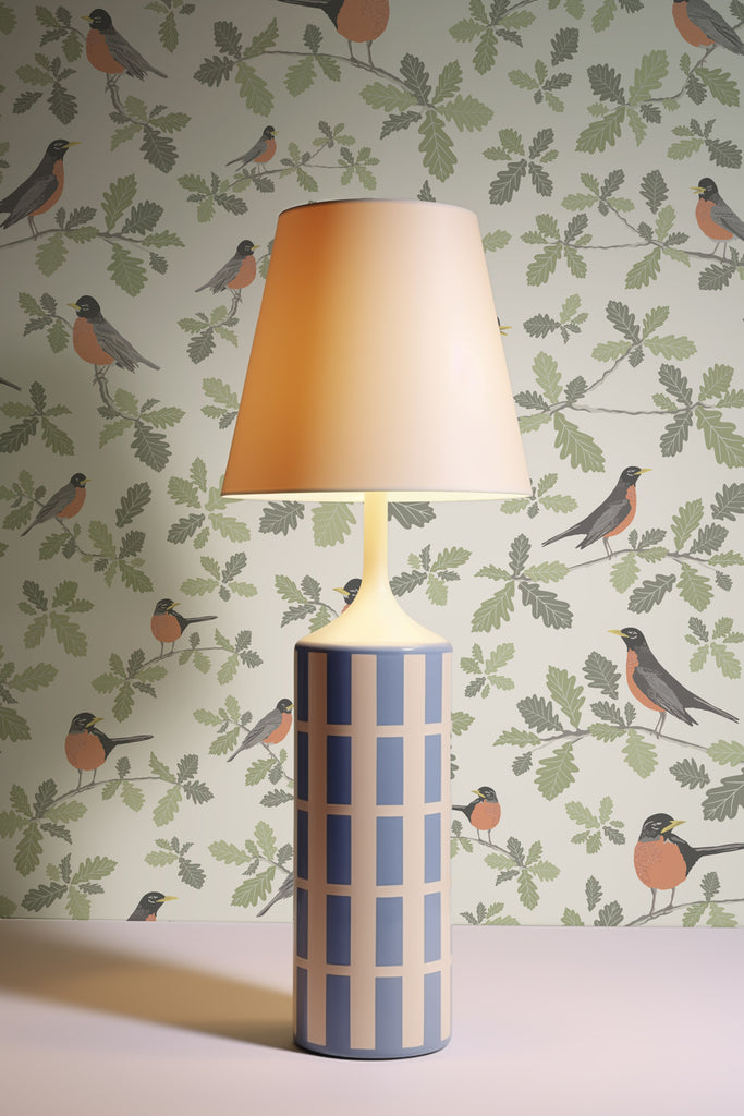 A sleek, modern lamp casts a warm glow on the Robin Parchment wallpaper, highlighting the playful bird motifs and soft foliage