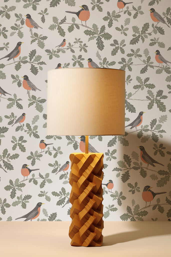 A chic, carved wooden lamp stands before the Robin Silvery Grey wallpaper, casting a warm glow on the lively robin scene, infusing the room with the essence of spring.